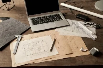 Ein Schreibtisch mit einem Laptop, Bauplänen und technischen Zeichengeräten.