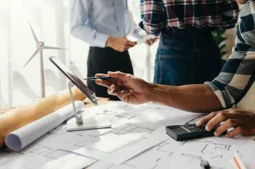 Eine Person arbeitet an einem Tisch mit Bauplänen und einem Tablet, im Hintergrund sind weitere Personen zu sehen.